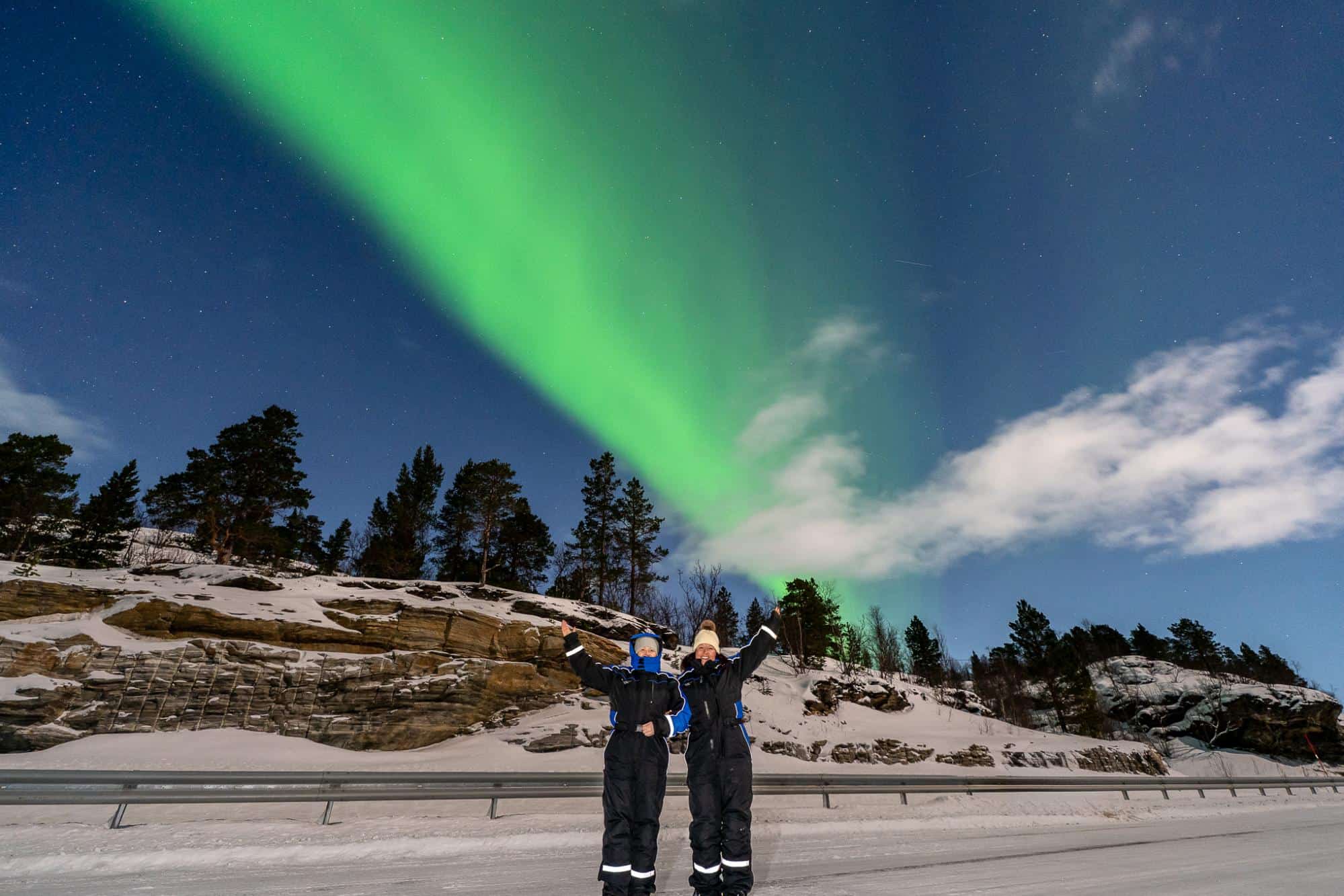 Two people in winter clothing stand with arms raised on a snowy road at night, with a vivid green aurora borealis lighting up the sky above snowy trees and rocky terrain.