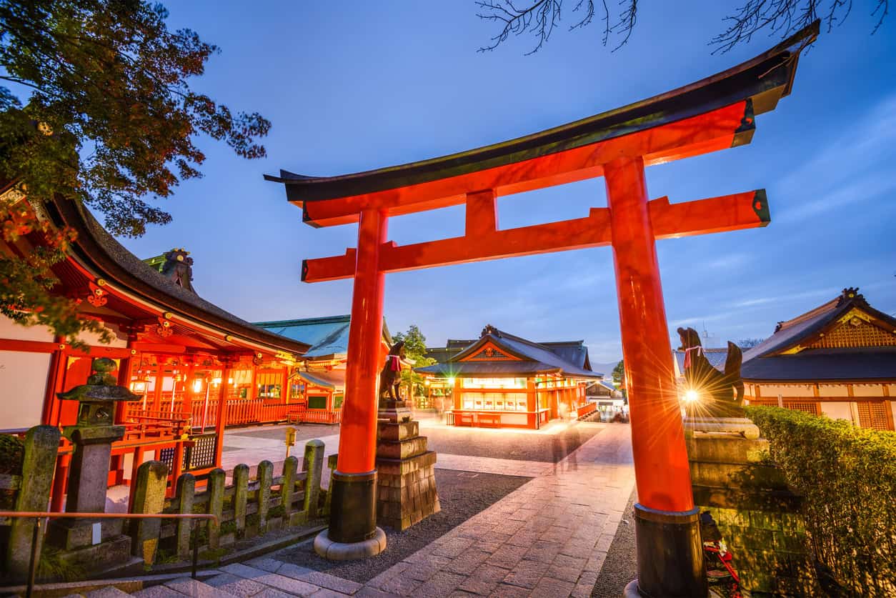 A large red torii gate stands at the entrance to a brightly lit Shinto shrine complex in Japan at dusk, traditional buildings and lanterns glowing—an enchanting scene near where you might arrange your Park Hyatt Kyoto booking.