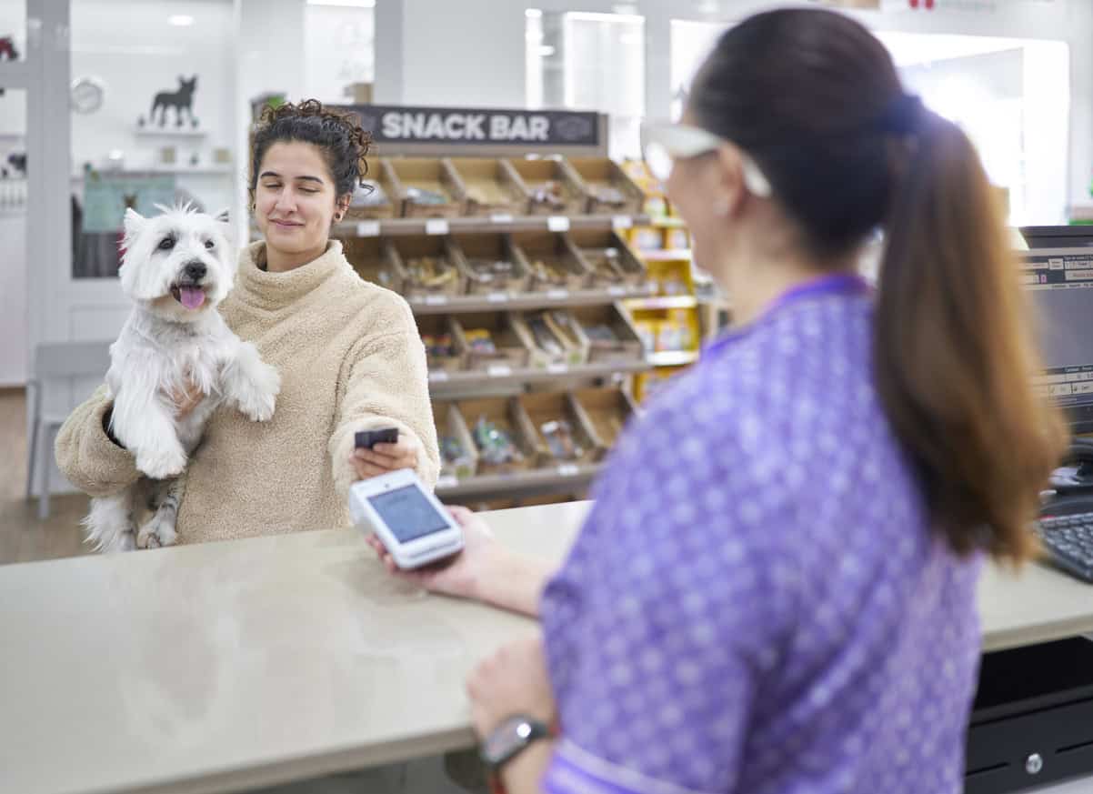 A woman holding a happy white dog pays at a counter, likely in a pet shop or vet clinic,—reminding us it’s possible to turn unexpected expenses into travel with the right planning.
