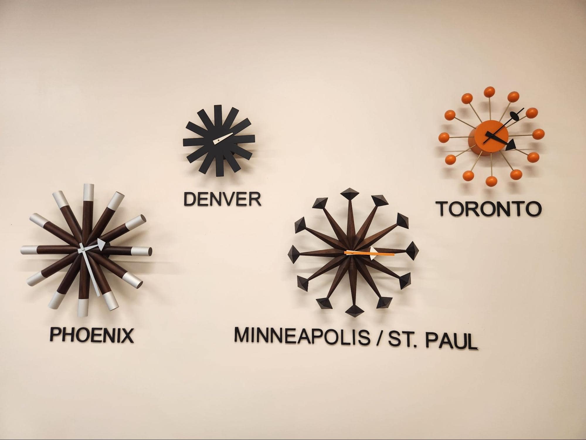 Four modern wall clocks above city names (Phoenix, Denver, Minneapolis/St. Paul, Toronto) each have unique decorative designs and display different times, mounted on a plain beige wall.