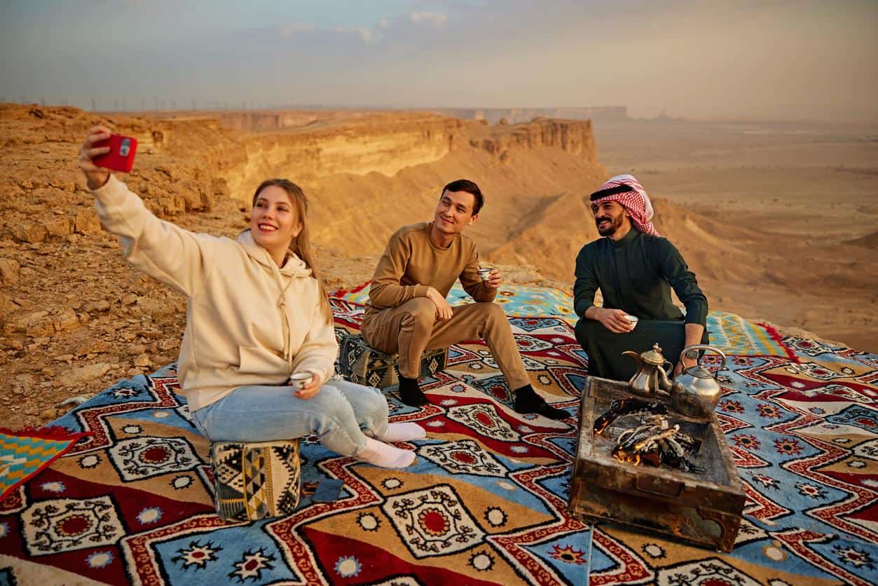Tourists enjoying traditional desert camping in Saudi Arabia with tea and scenic canyon views.