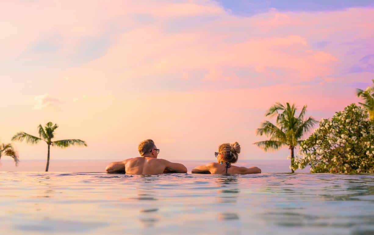 Couple relaxing in an infinity pool at sunset with palm trees and ocean in the background