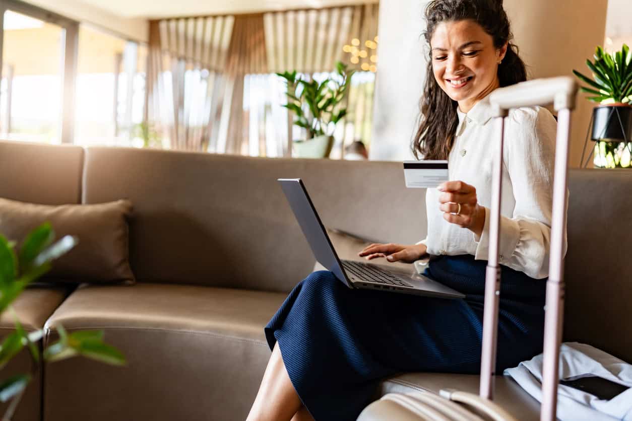 A woman sits on a sofa with a suitcase nearby, smiling while holding a credit card and using a laptop in a bright, modern lobby—exploring how to use onekeycash for seamless travel bookings.
