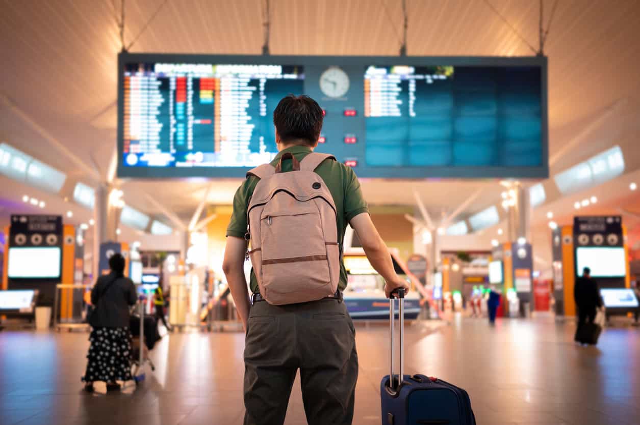 A person with a backpack and suitcase stands in an airport terminal, gazing up at a large digital departures board, perhaps planning to use their Amex annual credits for travel perks.