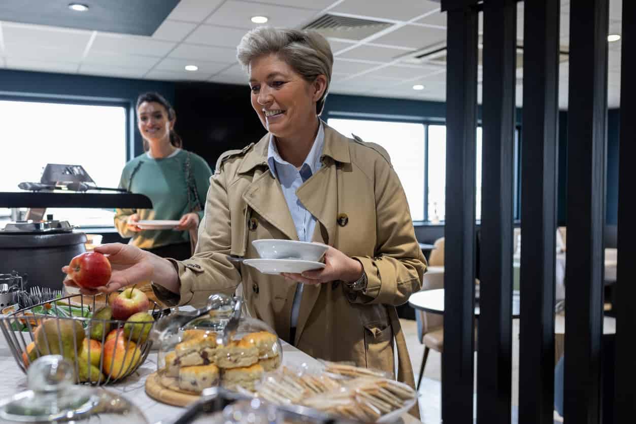Smiling woman in an airport lounge buffet selecting an apple while holding a bowl, with fresh fruits and pastries on display.