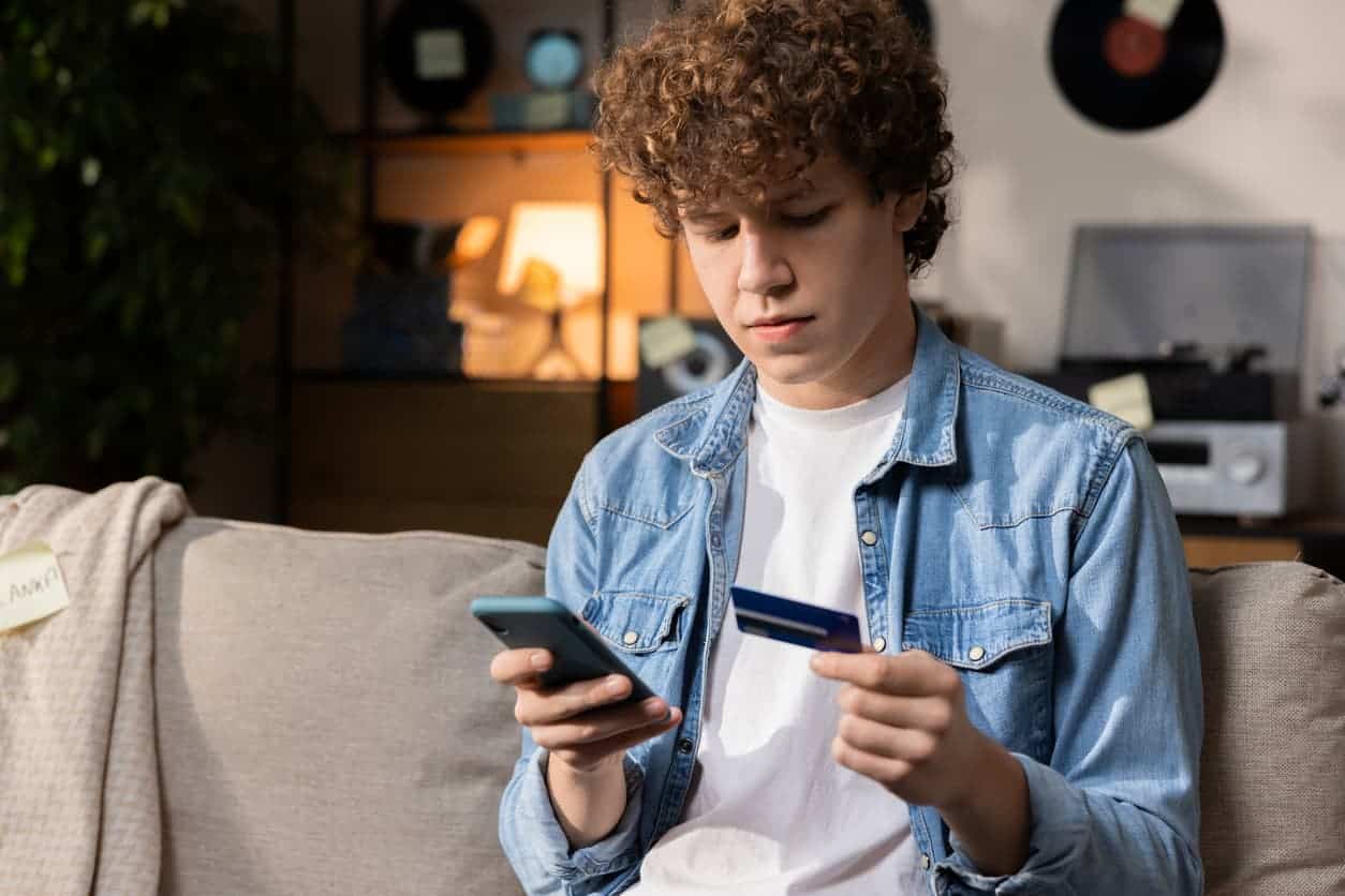 Teenage boy sitting on a couch making an online purchase using a credit card and smartphone