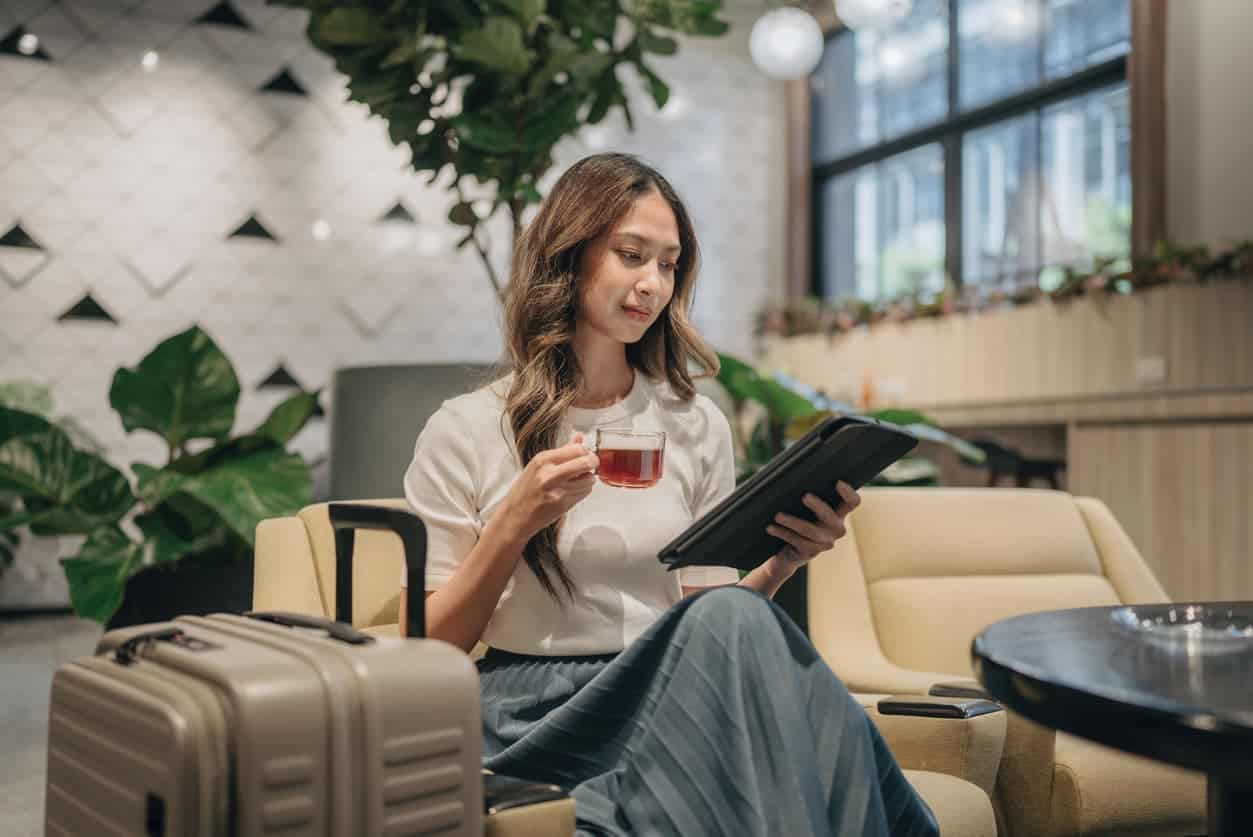 Female traveler sitting in an airport lounge with luggage, drinking tea and reading a tablet.