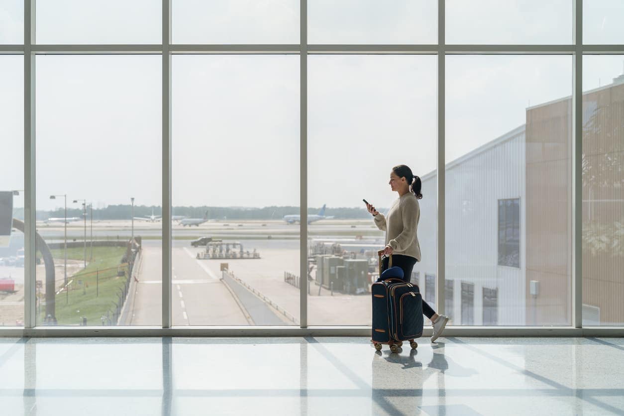 Woman walking through airport terminal with rolling suitcase while checking phone