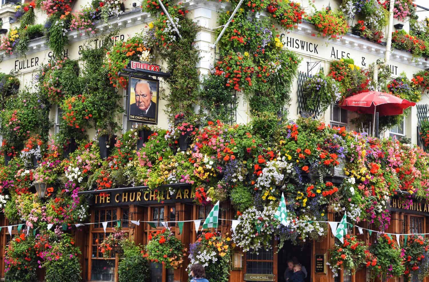 A traditional British pub called The Churchill Arms is decorated with a dense, colorful array of flowers covering the exterior. Patrons are visible at the entrance; signs and a portrait of Winston Churchill are displayed.