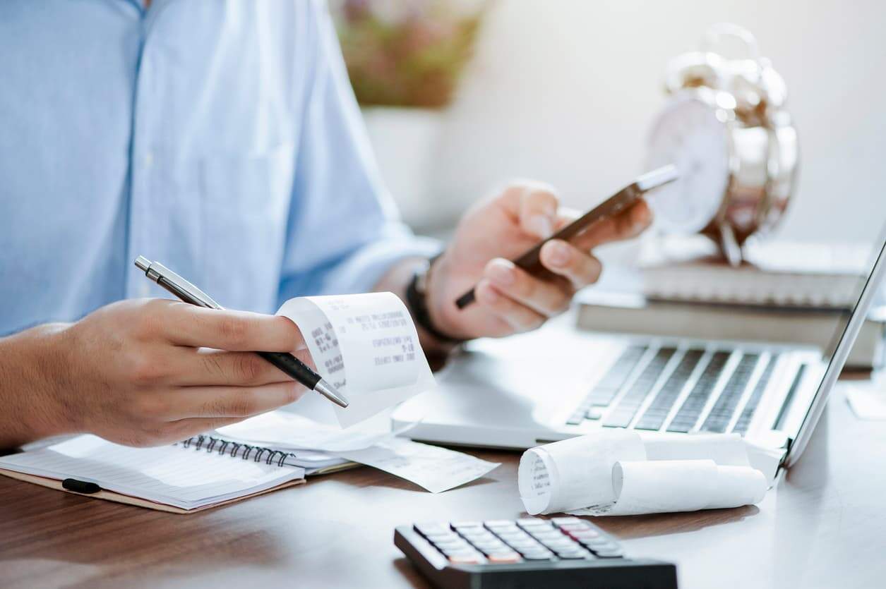 Man calculating tax payments using receipts, smartphone, and laptop at a desk.