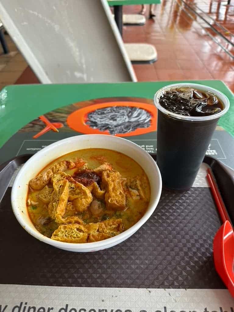 Bowl of Singapore laksa noodle soup with tofu puffs served alongside a cup of iced coffee