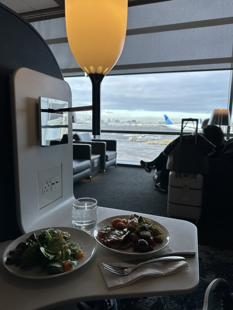 Quiet corner at United Polaris Lounge EWR with food and a view of planes on the runway through large windows.