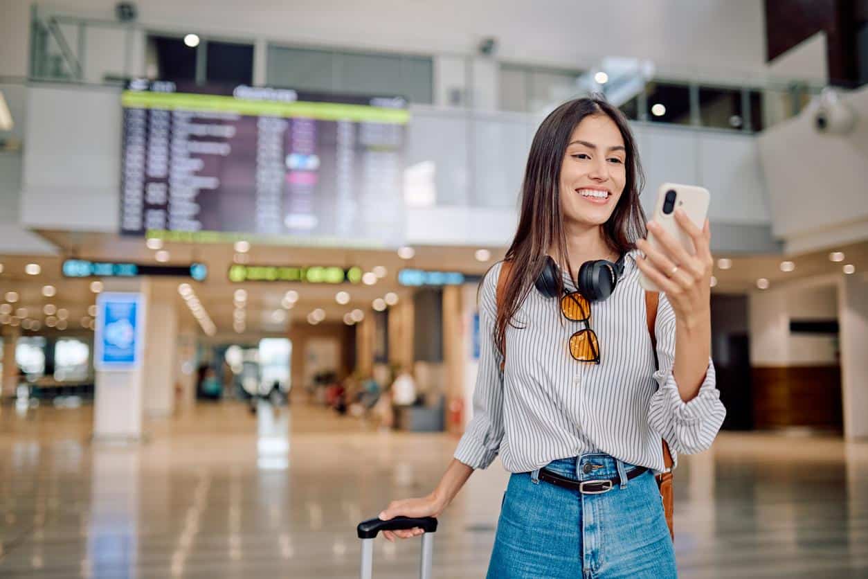 Woman at airport smiling while looking at her phone and holding luggage.