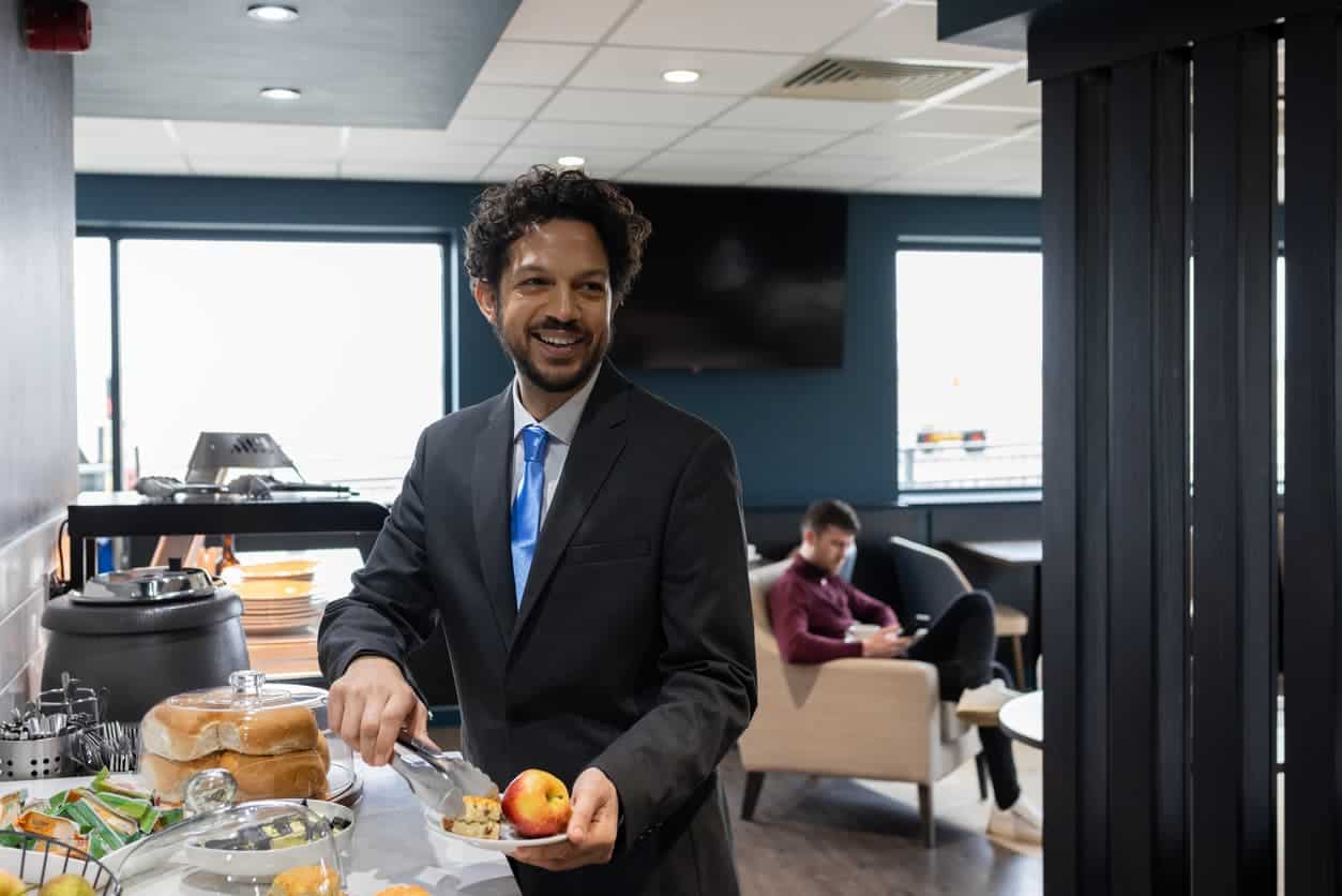Man in a suit serving himself food from a breakfast buffet inside an airport lounge.