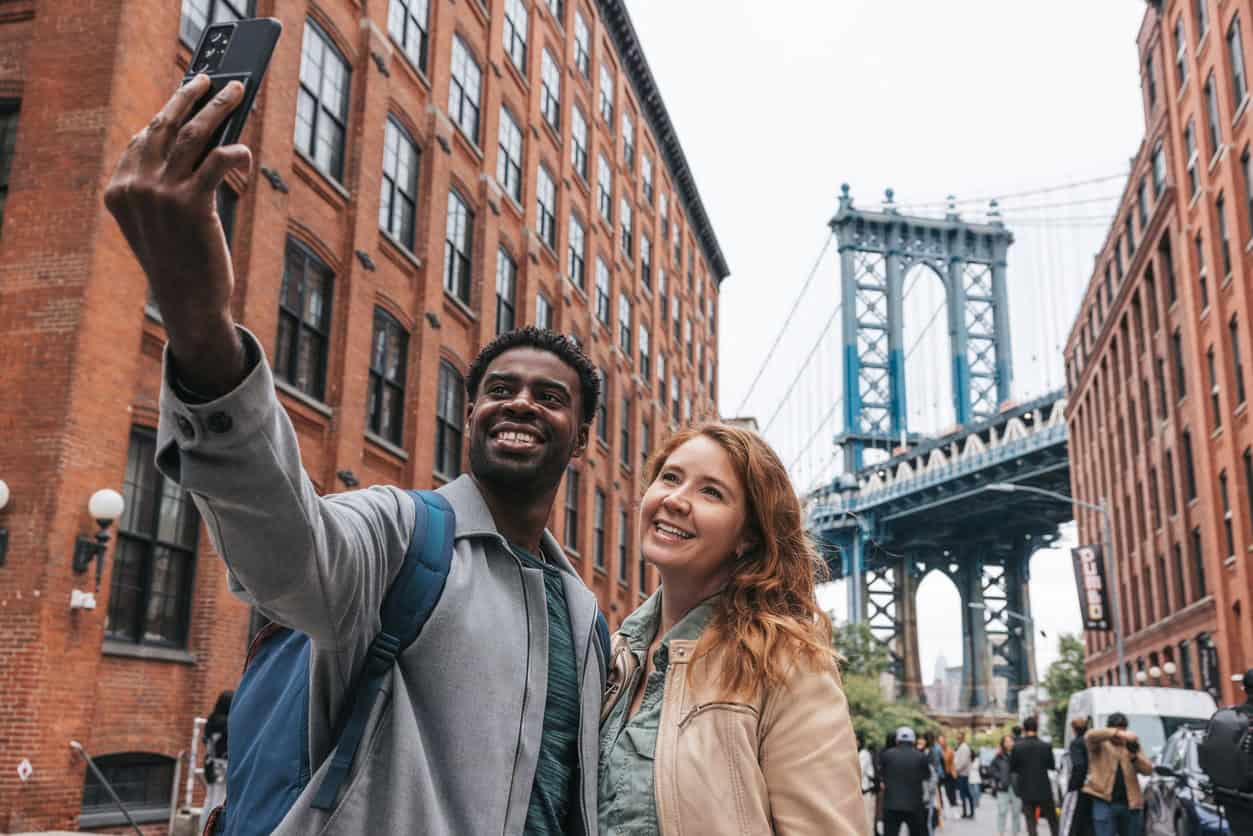 Two travelers taking a selfie in the DUMBO neighborhood of Brooklyn with the Manhattan Bridge in the background.
