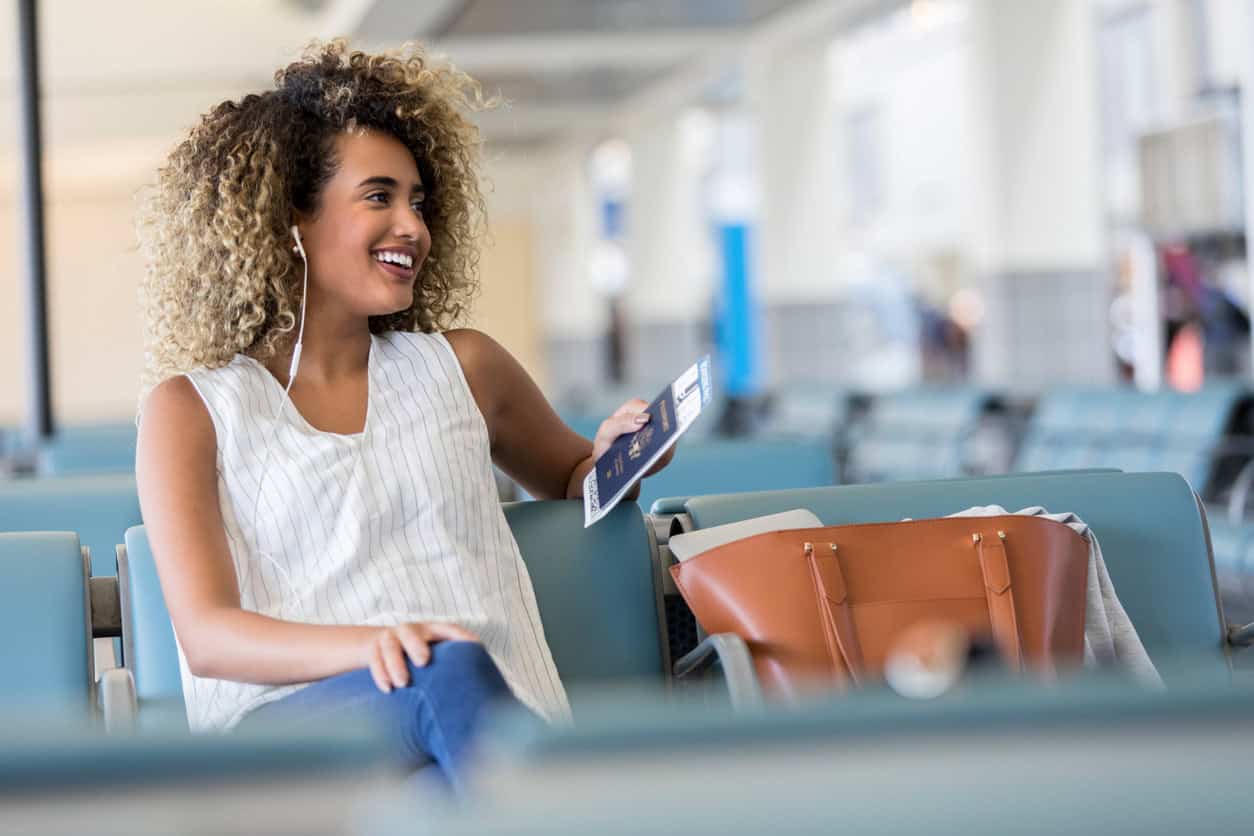 Smiling woman sitting in an airport terminal holding a passport and boarding pass with her bag beside her.