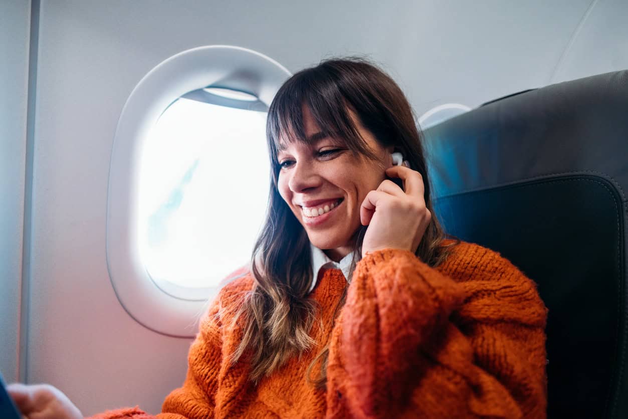 woman smiling while listening to audio on a flight, seated by the airplane window