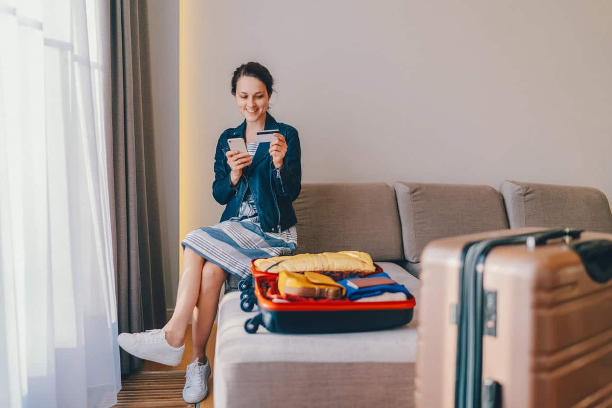 Woman sitting in a hotel room using her phone and credit card while packing luggage for a trip.