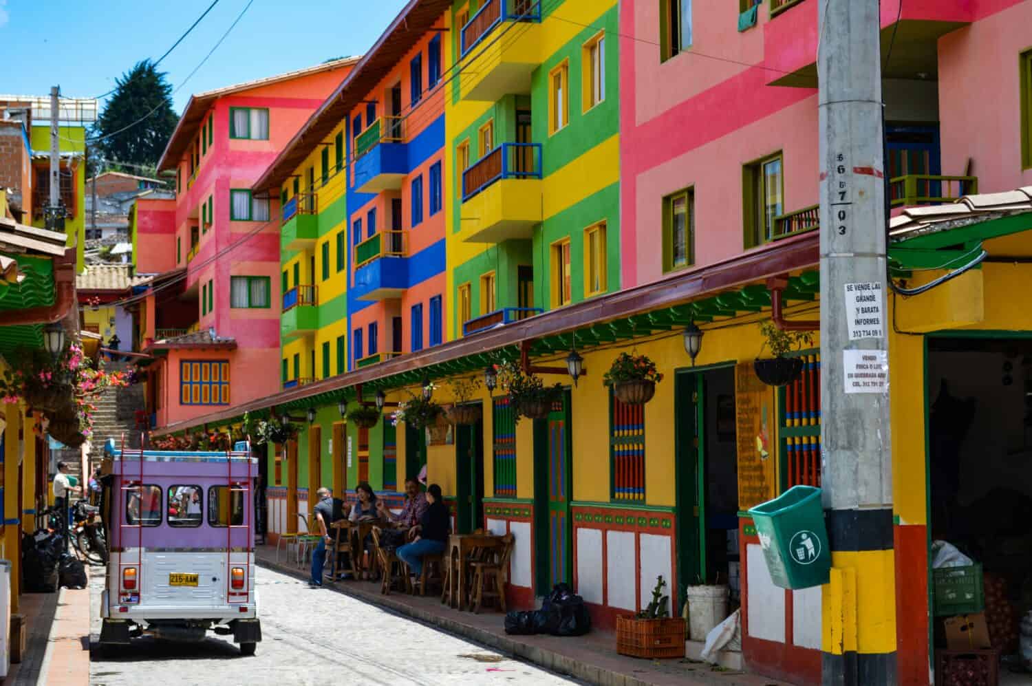A narrow street lined with bright, multicolored buildings; people sit at tables outside a café, and a purple vehicle drives past. Hanging plants and vivid paint create a lively atmosphere in Colombia.