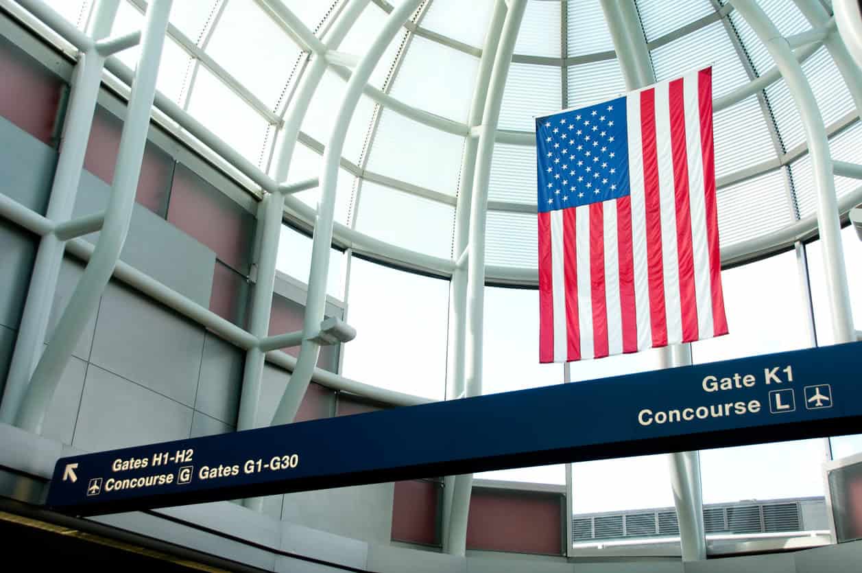 Airport terminal interior with overhead concourse and gate signage beneath a large American flag.