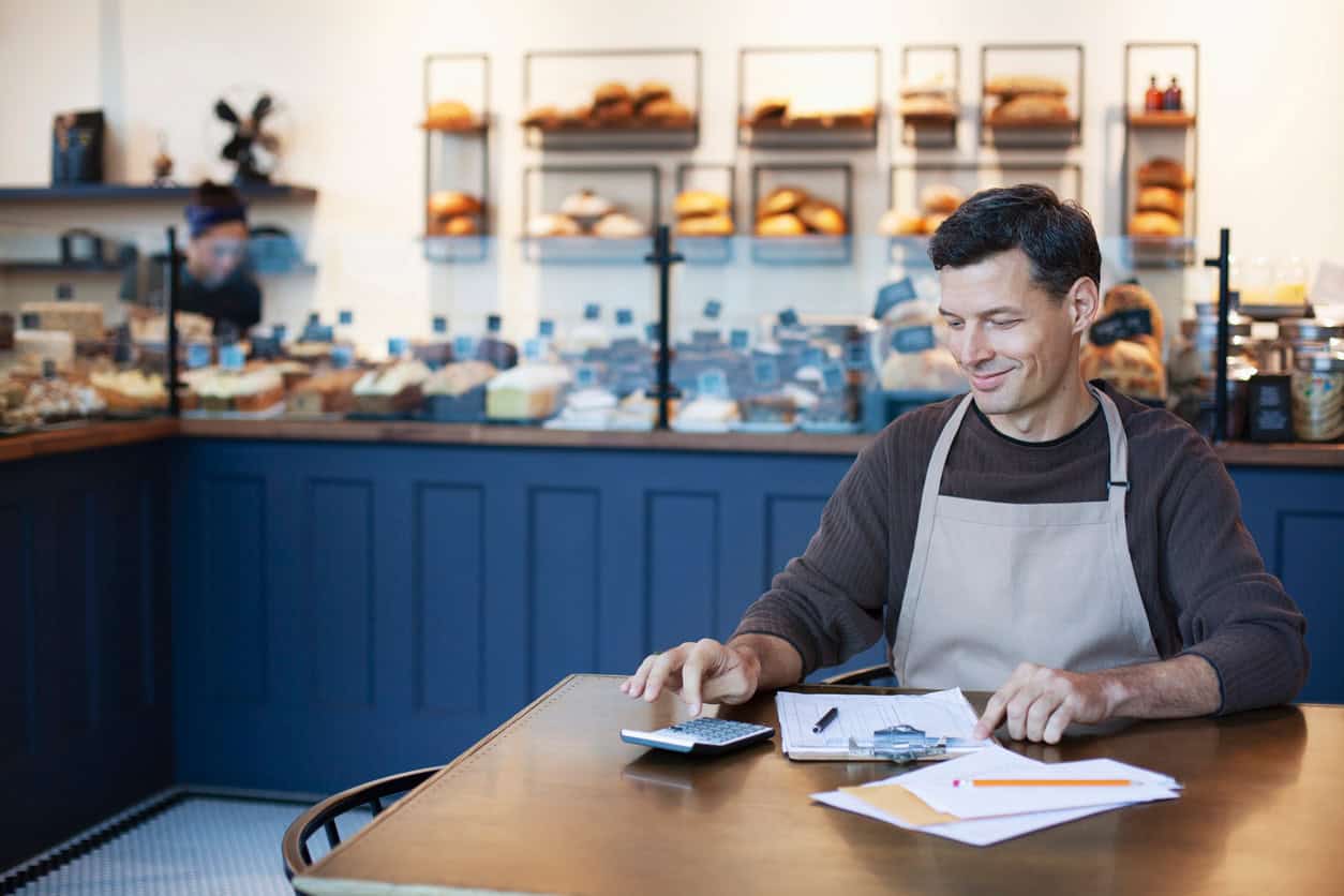 Bakery owner in an apron using a calculator to review business expenses at a shop counter