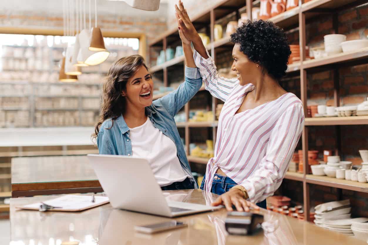 Two business owners celebrating a successful sale in a retail shop, using a laptop and card reader with a business cashback card