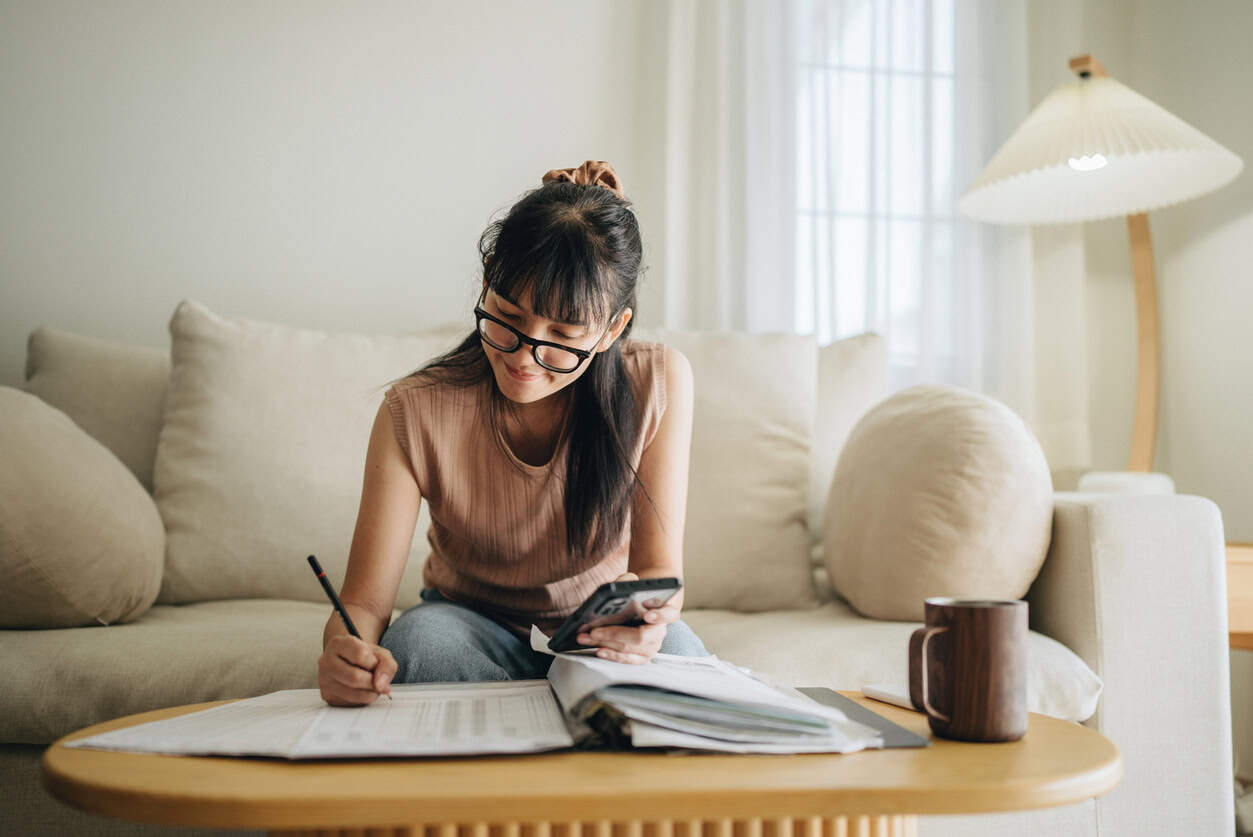 Business owner at home reviewing paperwork and calculating expenses with a smartphone and notebook