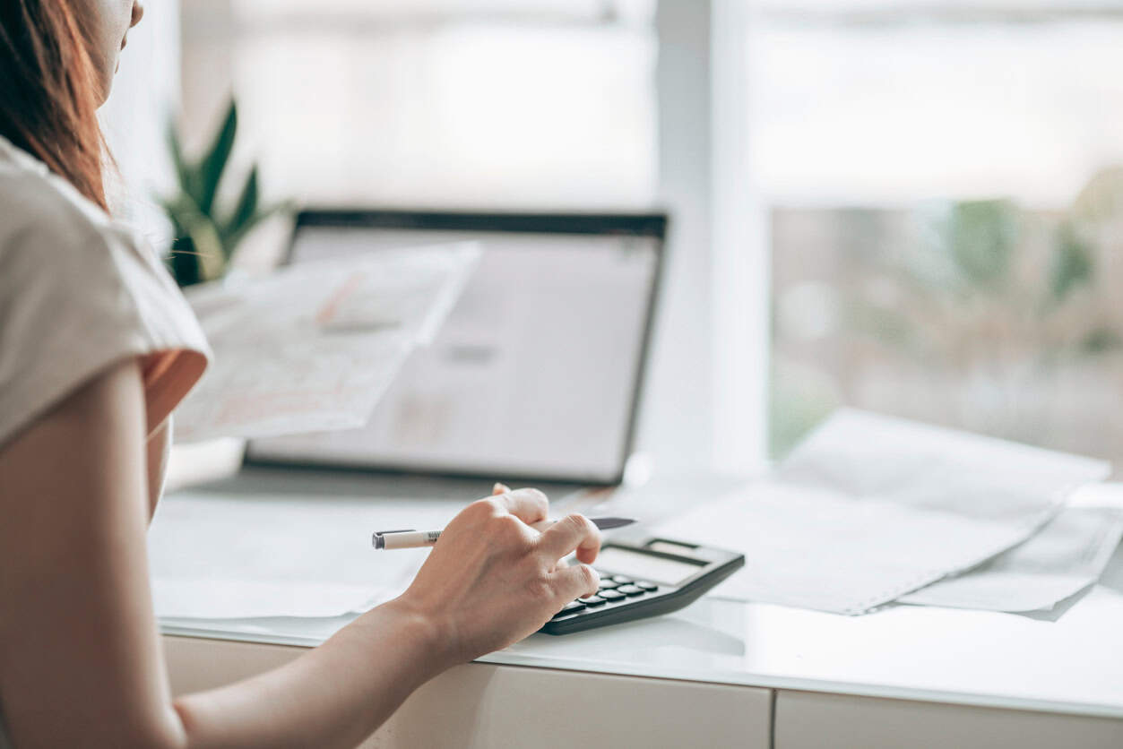 Person calculating business expenses at a desk using a calculator and reviewing paperwork near a window