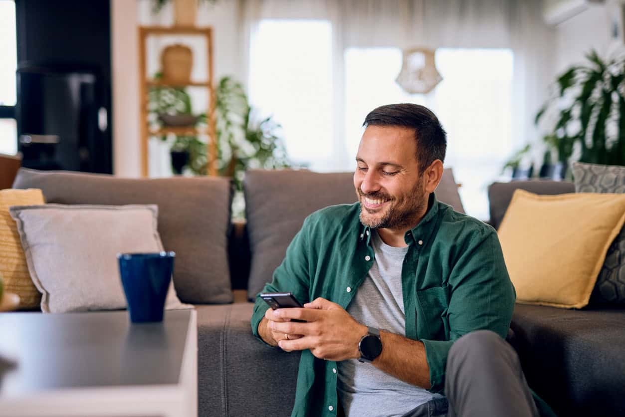 Man sitting on a sofa at home, smiling while using his smartphone in a bright living room