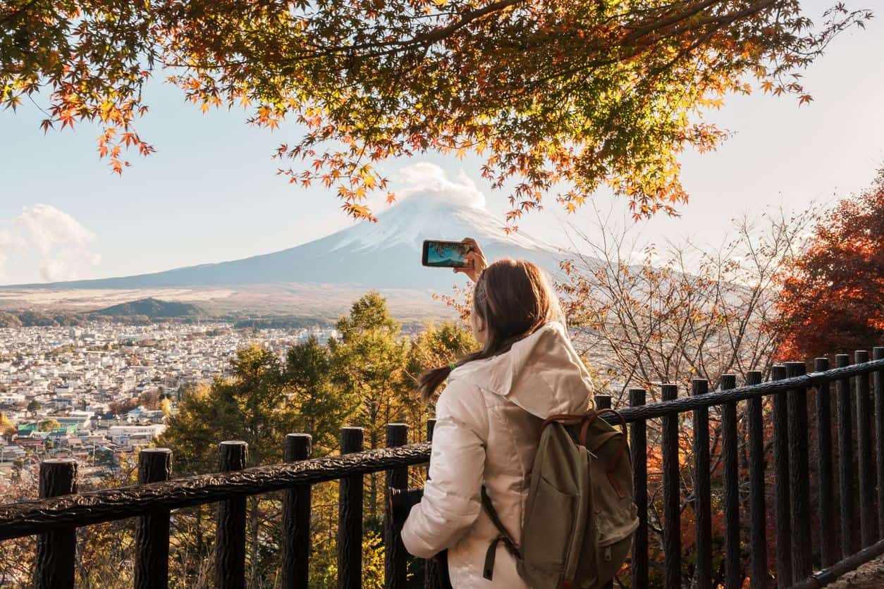 Traveler taking a photo of Mount Fuji during autumn with colorful maple leaves overlooking a Japanese town
