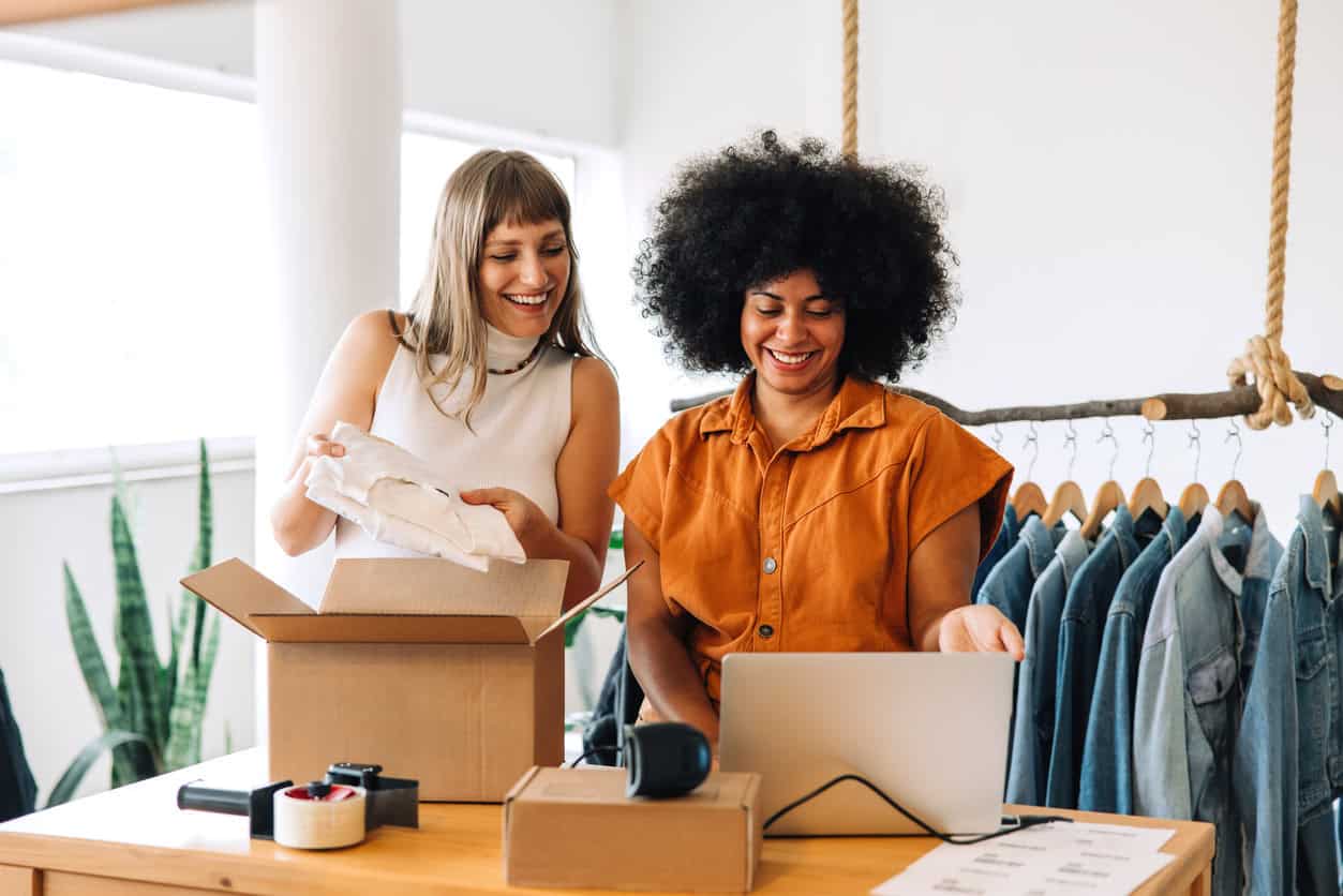 Two small business owners packing online orders at a workspace with clothing racks and a laptop used to manage sales and shipping