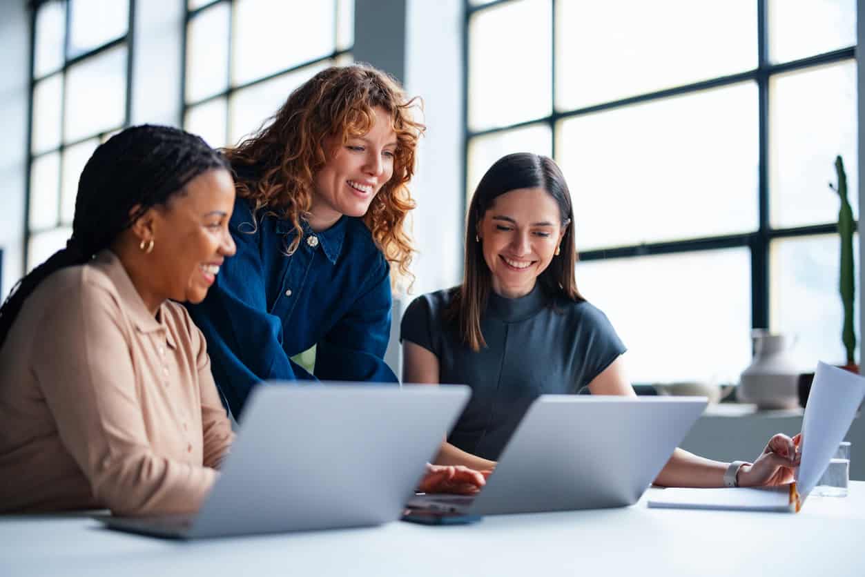 Three colleagues collaborating at a desk while reviewing documents on laptops