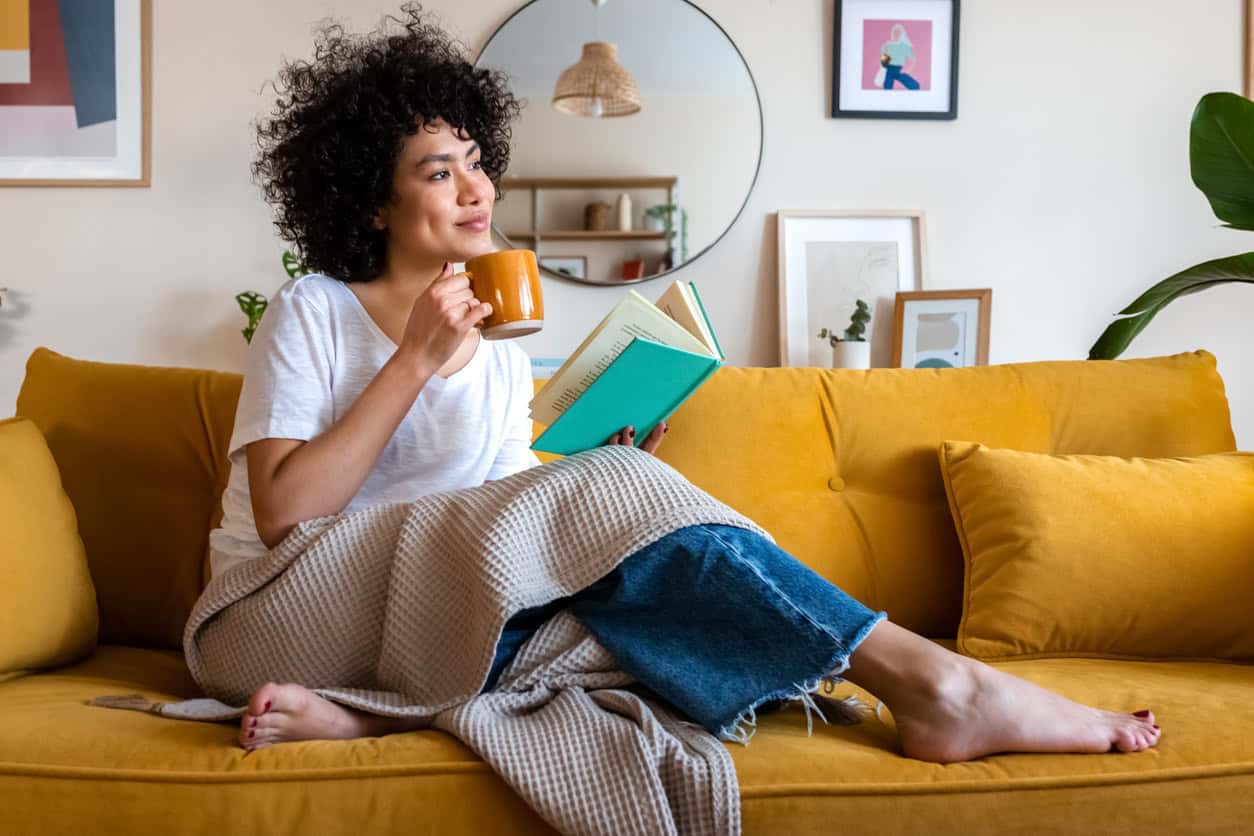Woman relaxing on a yellow sofa, reading a book and holding a mug in a cozy living room