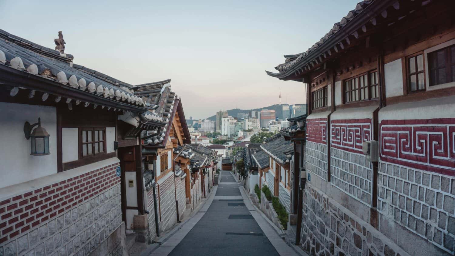 A narrow street lined with traditional Korean houses leads down toward a modern city skyline, blending old and new architecture under a clear sky.