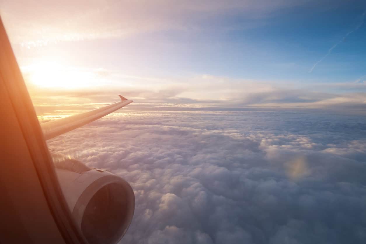 View from airplane window showing wing and engine above clouds during sunrise flight