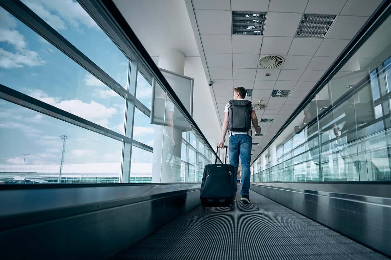 Traveler walking through a modern airport terminal on a moving walkway while pulling a carry-on suitcase.