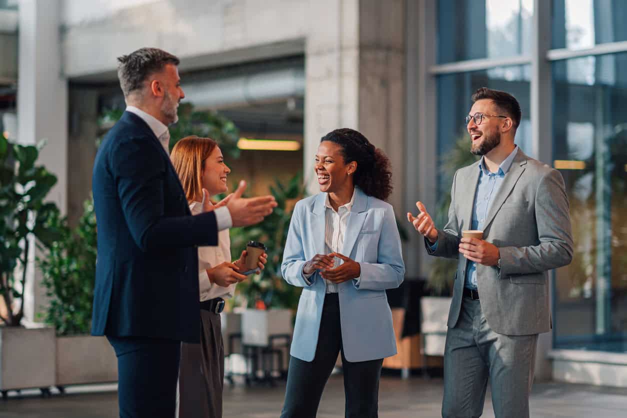 Group of business professionals talking and laughing together in office lobby during networking break with coffee cups