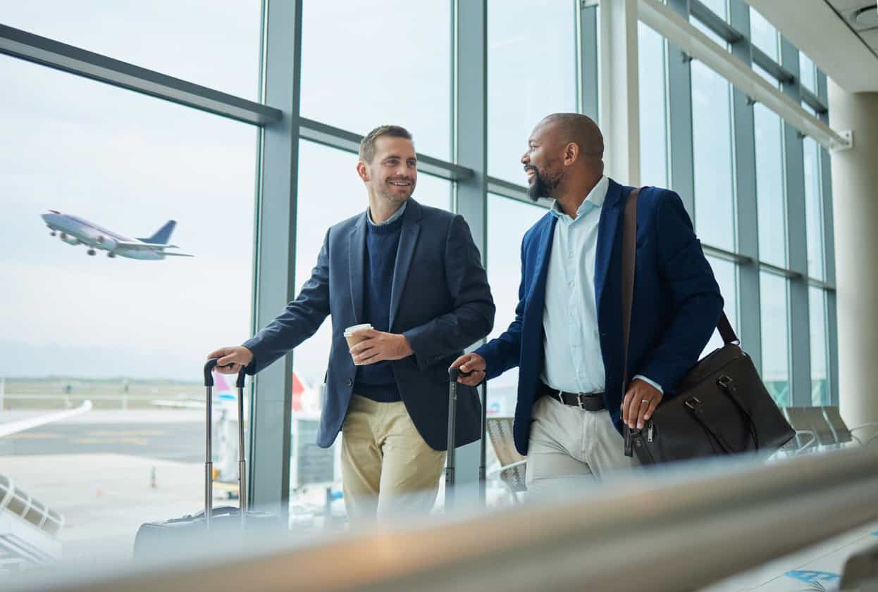 Two business travelers walking through airport terminal with rolling luggage and airplane visible outside window