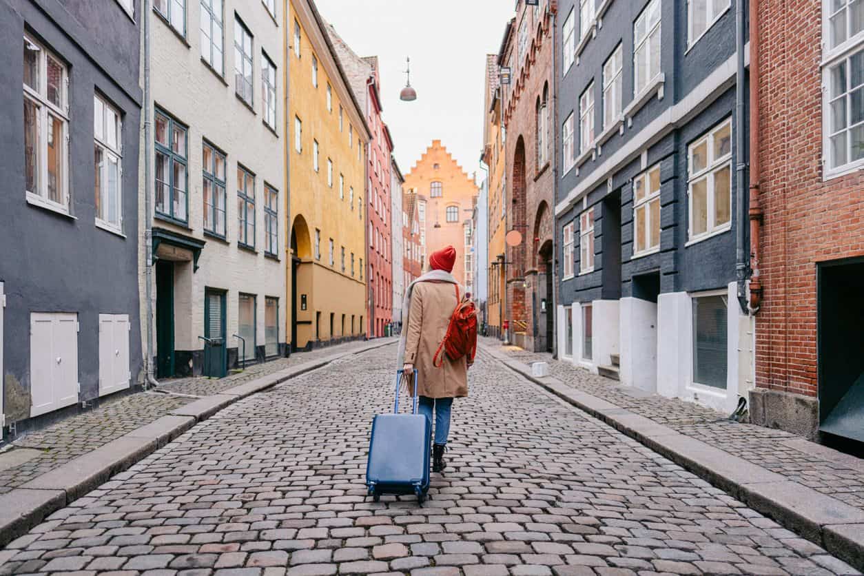 Traveler pulling a rolling suitcase along a cobblestone street lined with colorful historic buildings in Denmark, likely Copenhagen