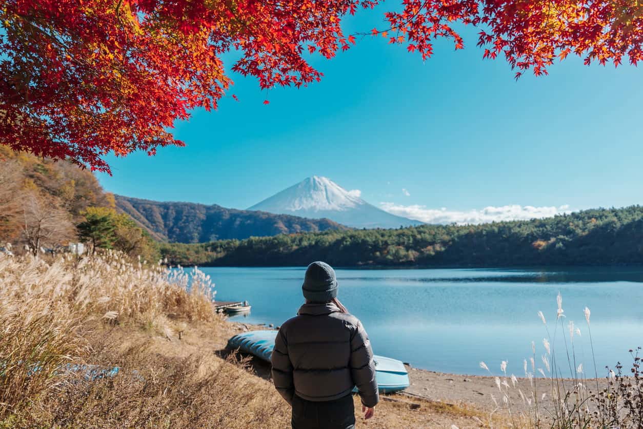 Traveler looking at Mount Fuji across a calm lake framed by red autumn leaves in Japan