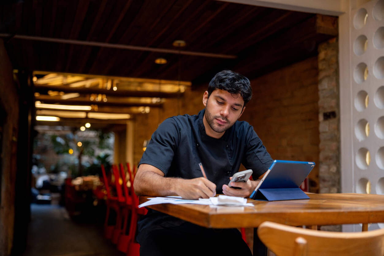 Small business owner reviewing receipts and calculating expenses on phone and tablet at restaurant table for tax filing
