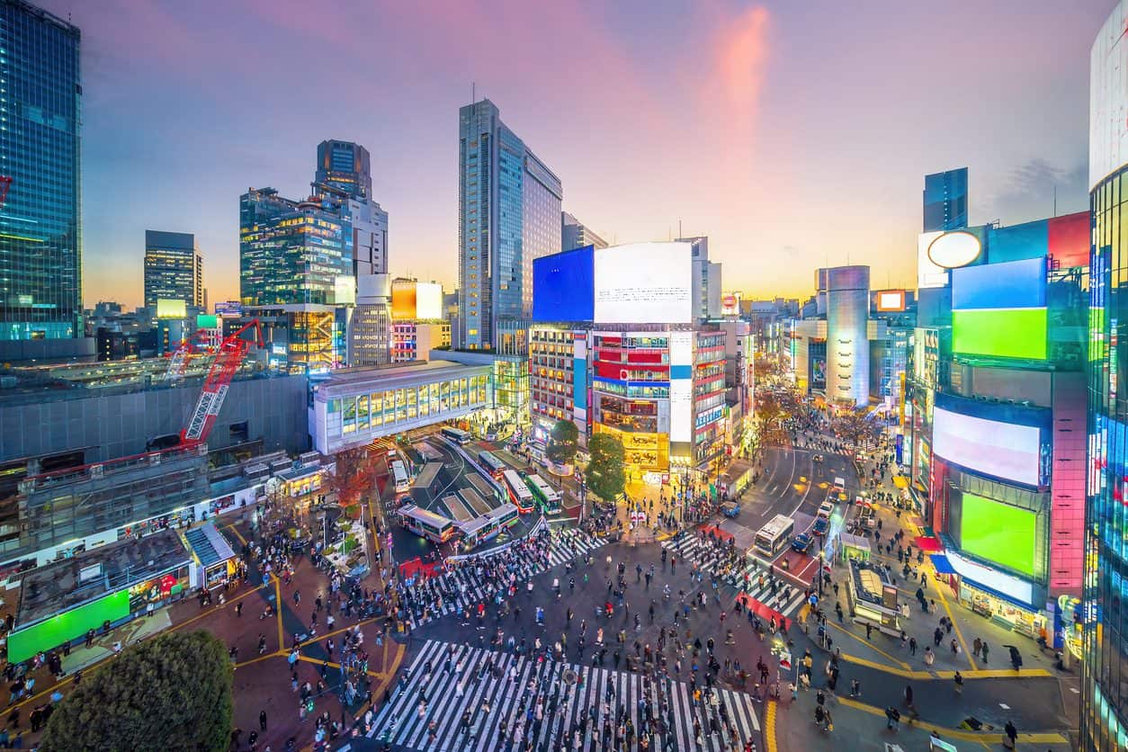 Aerial view of Shibuya Crossing in Tokyo at dusk with illuminated billboards, busy crosswalks, and crowds moving through the intersection