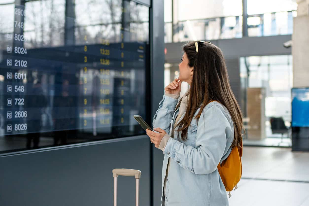 Female traveler with backpack and suitcase checking airport departure board while holding phone