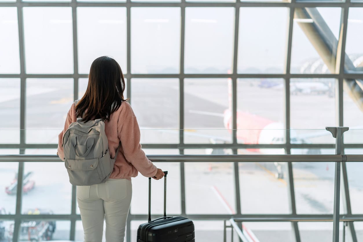 Traveler with backpack and rolling suitcase looking out airport window at airplane before departure