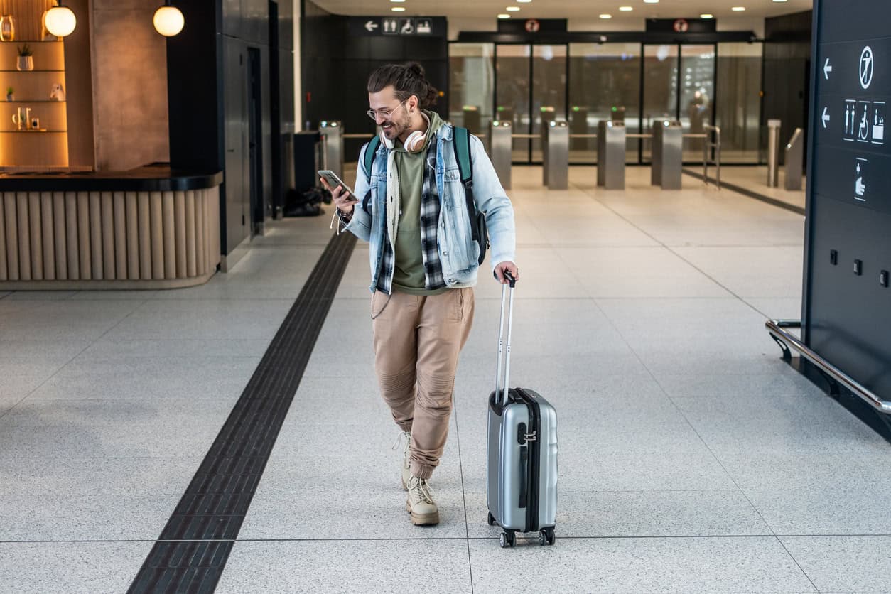 Traveler walking through airport terminal pulling carry-on suitcase while checking phone for flight details