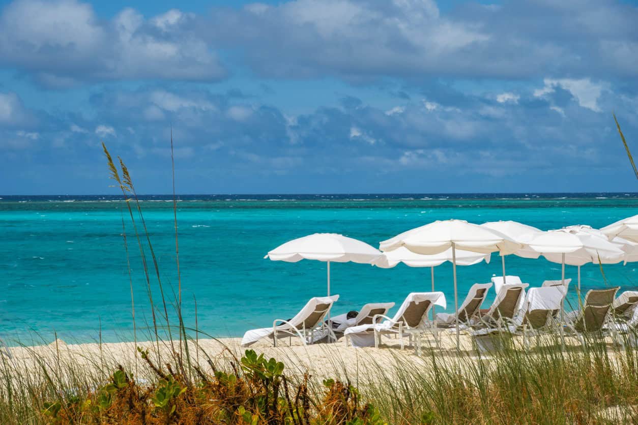 White sand beach in Turks and Caicos with turquoise ocean water, beach loungers, and white umbrellas under a partly cloudy sky