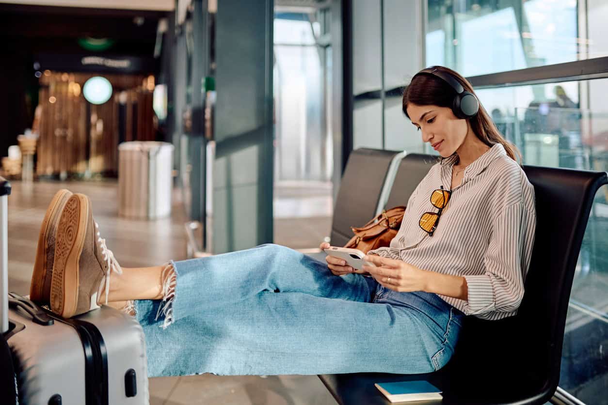 Woman wearing headphones sitting at an airport gate using her smartphone with carry-on luggage nearby