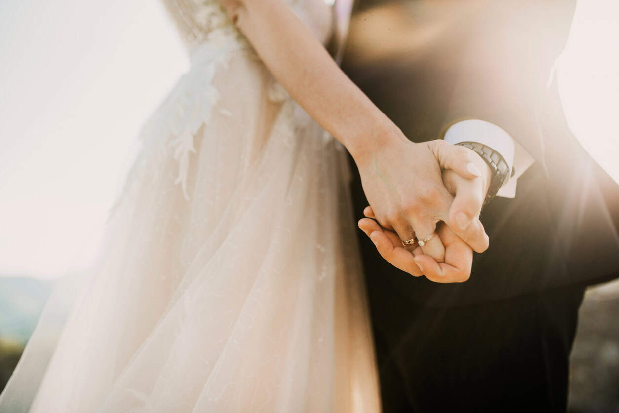Close-up of bride and groom holding hands with wedding rings visible during outdoor wedding ceremony