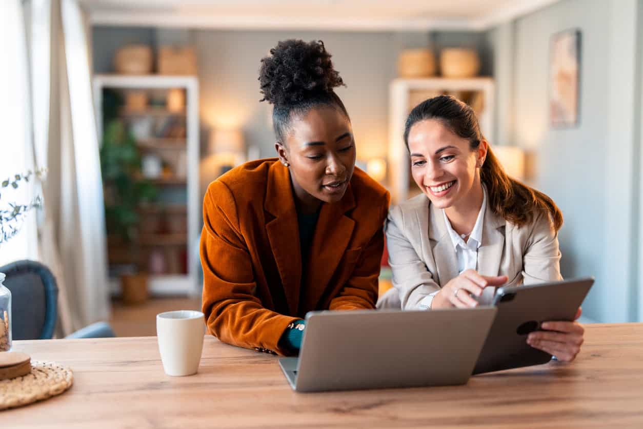 Two business owners collaborating on laptop and tablet in a home office workspace, discussing work and smiling
