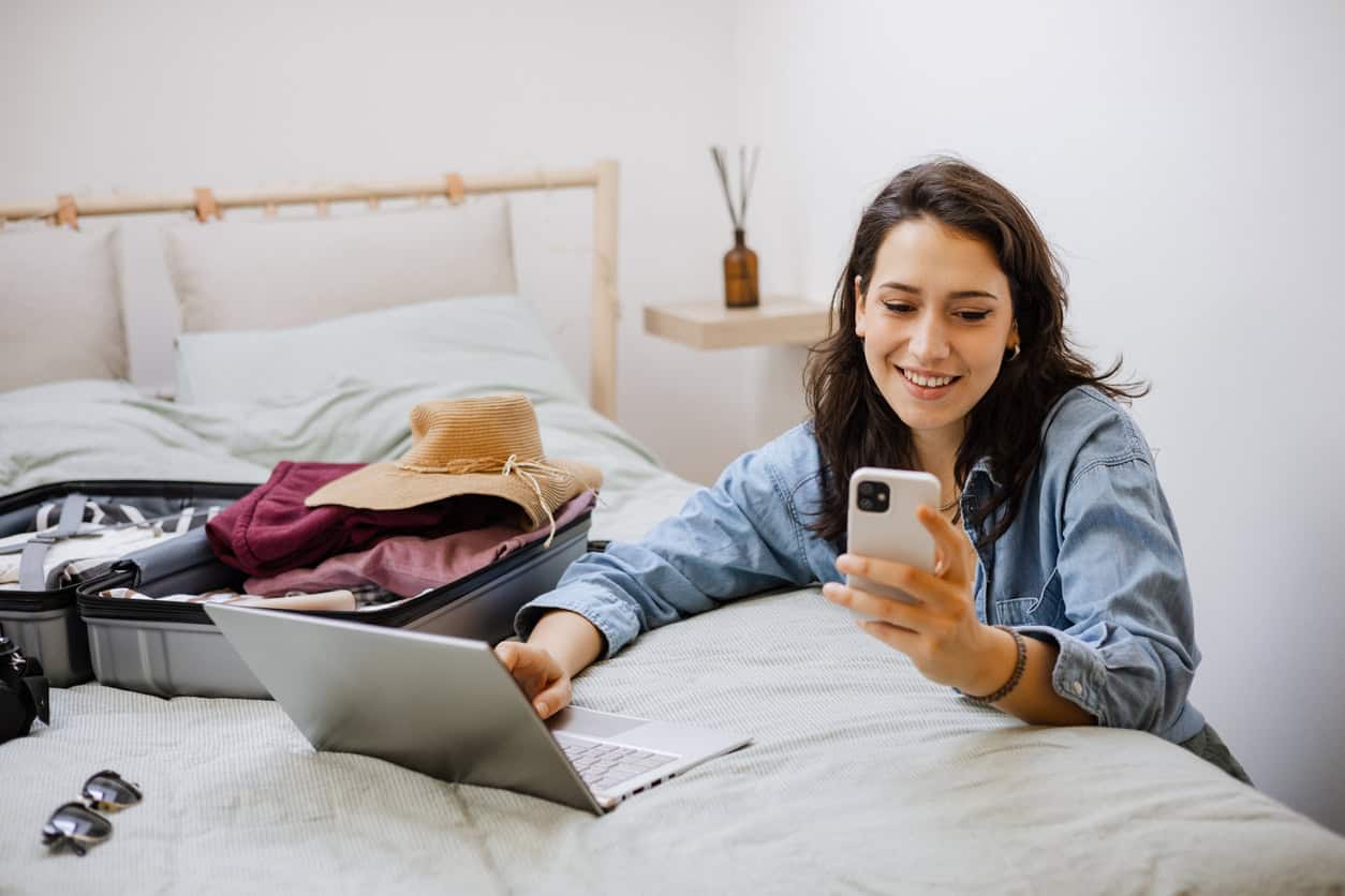 Woman using phone and laptop while packing suitcase and sharing travel plans and redemptions